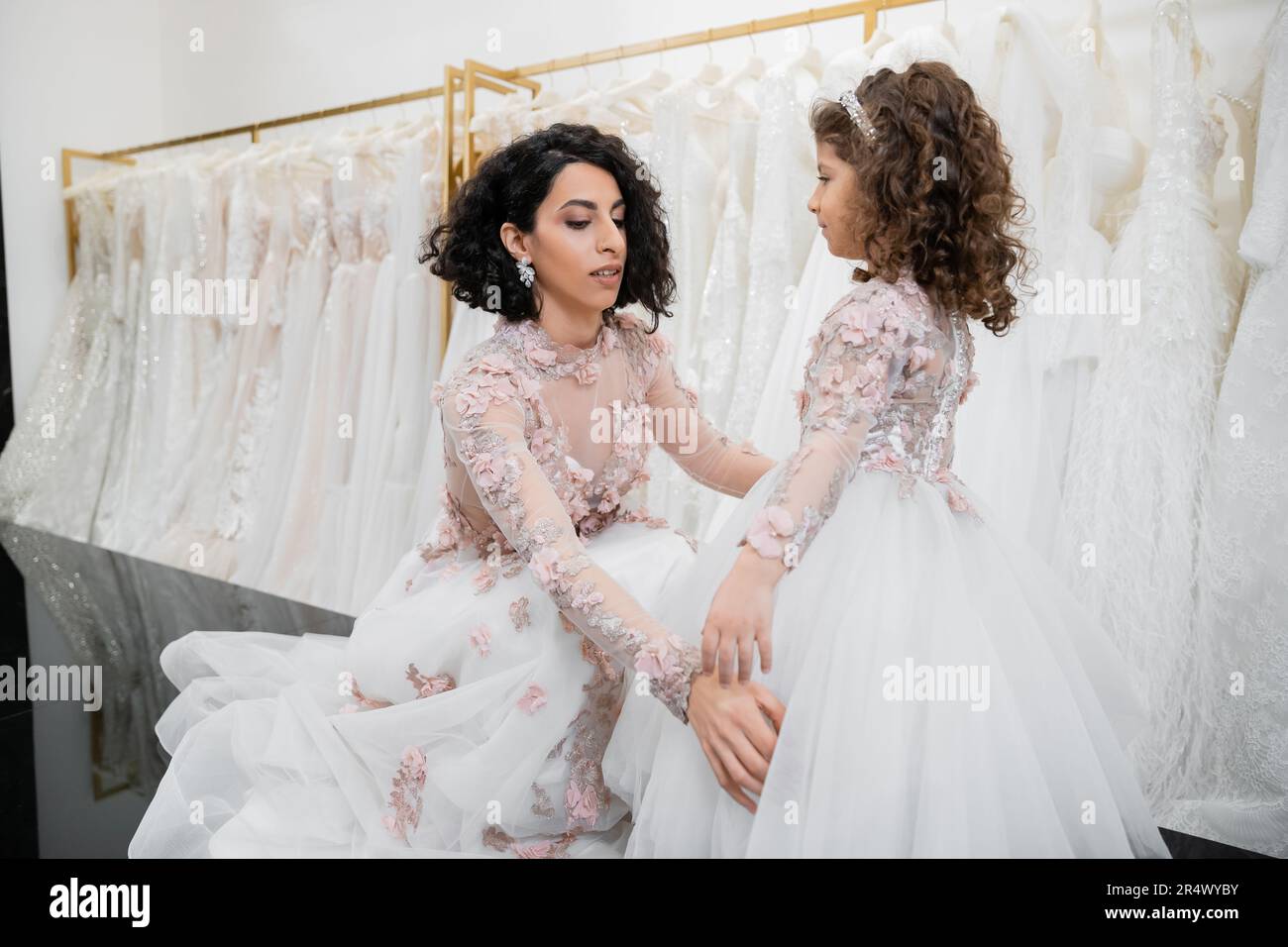 special moment, brunette middle eastern bride in floral wedding gown adjusting skirt of her little daughter`s attire in bridal salon around white tull Stock Photo